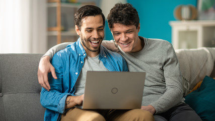 Adorable Male Gay Couple Spend Time at Home. They Sit on a Sofa and Use the Laptop. They Watch Funny Online Videos. Partner Puts His Hand Around His Lover. Room Has Modern Interior.