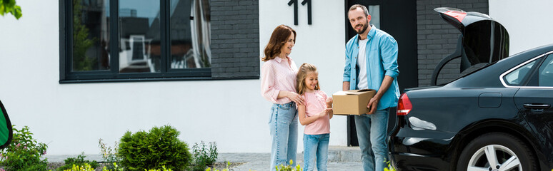 Fototapeta na wymiar panoramic shot of cheerful family moving into modern house while standing near car