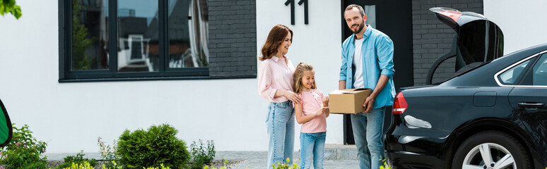 panoramic shot of cheerful family moving into modern house while standing near car