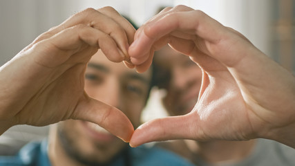 Portrait of a Cute Male Queer Couple at Home Sitting on a Sofaand Making a Heart Symbol with Their...
