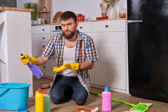 Caucasian young bearded man sits on the floor of his kitchen and tries to clean it using all of his detergents, rags, washcloths and brushes