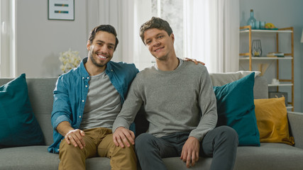 Cute Attractive Male Gay Couple Sit Together on a Sofa at Home. Boyfriend Puts His Hand on Fiance's. They are Happy and Smiling. They are Casually Dressed and Their Room Has Modern Interior.