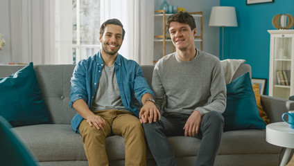 Cute Attractive Male Gay Couple Sit Together on a Sofa at Home. Boyfriend Puts His Hand on Fiance's. They are Happy and Smiling. They are Casually Dressed and Their Room Has Modern Interior.