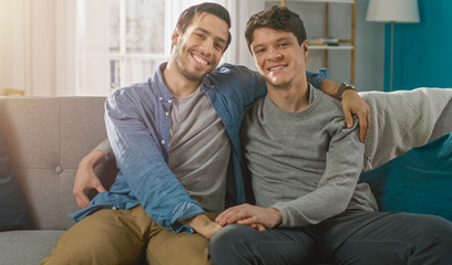 Cute Attractive Male Gay Couple Sit Together on a Sofa at Home. Boyfriend Puts His Hand on Partner's and They Hug. They are Happy and Smiling. They are Casually Dressed and Room Has Modern Interior.