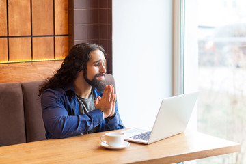 Please! Portrait of worry handsome young adult man freelancer in casual style sitting in cafe and talking with his friend in laptop,holding hand like pray and apologies. Indoor, lifestyle concept