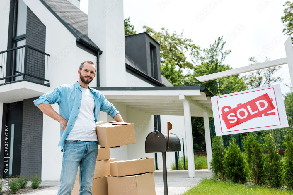 Wall mural handsome man standing with hand on hip and holding box near house and board with sold letters