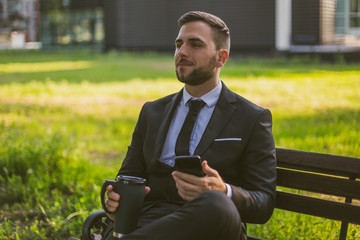 Obraz premium Elegant businessman is drinking coffee and using phone at the park.