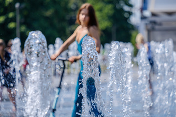 A girl in a tight-fitting suit of heavenly color riding a scooter in the middle of the fountain...