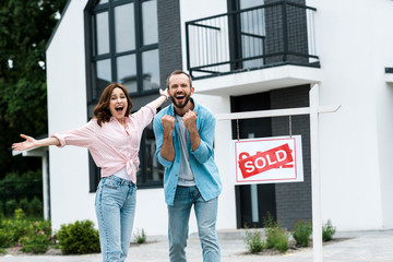 cheerful man and woman celebrating near house and board with sold letters - obrazy, fototapety, plakaty