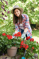 Beautiful smiling Caucasian brunette in working clothes and with hat pruning flowers in her backyard.