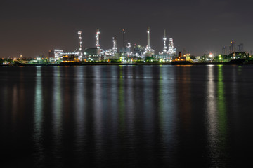 Night photo crude oil refinery plant and many chimney with petrochemical tanker or cargo ship at coast of the river with colorful bright light from lamp reflect on water at thailand