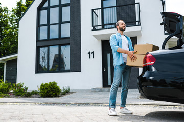 happy man holding boxes near car and modern house