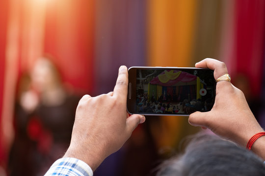 man holding mobile phones and shoot video on them. bright colored background of Indian concert in blur. the sharpness on the hand with phone