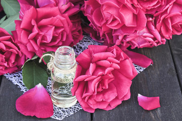 Glass bottle with rose essential oil and red roses on a dark wooden background.