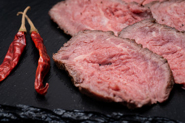 Sliced Grass Fed Juicy Corn Roast Beef garnished with dried Red Chile De Arbol Pepper on black natural stone background.