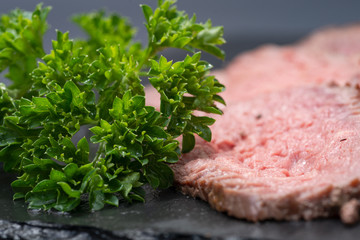 Sliced Grass Fed Juicy Corn Roast Beef garnished with Fresh Curly Parsley on black natural stone background.