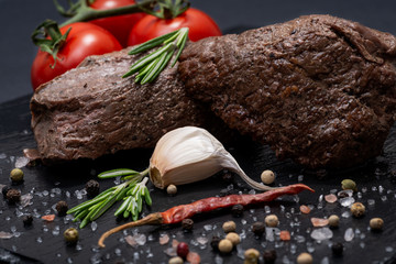 Grass Fed Corn Roast Beef garnished with Tomatoes, Fresh Rosemary, dried Red Chile Pepper, Garlic and Peppercorns on natural black stone background.