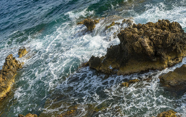 Summer cliff next to the Mediterranean Sea, strong waves break with the rocks and leave blue and turquoise colors along with the foam of the sea.