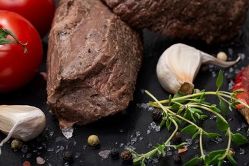 Grass Fed Corn Roast Beef garnished with Tomatoes, Fresh Thyme, dried Red Chile Pepper, Garlic and Peppercorns on natural black stone background.