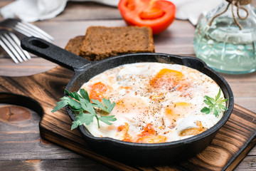 Homemade breakfast of shakshuka fried eggs with tomatoes and herbs in a pan on a cutting board on a wooden table