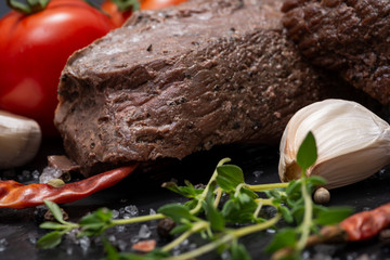 Grass Fed Corn Roast Beef garnished with Tomatoes, Fresh Thyme, dried Red Chile Pepper, Garlic and Peppercorns on natural black stone background.