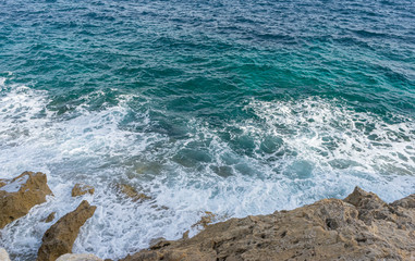 Fototapeta na wymiar Summer cliff next to the Mediterranean Sea, strong waves break with the rocks and leave blue and turquoise colors along with the foam of the sea.
