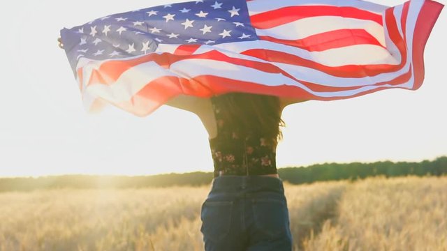 American teenager girl waving an American USA Stars and Stripes flag in a wheat field at sunset