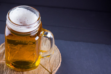 Beer with foam in mason jar on a dark background. A glass of light beer on a wooden stand. Low alcohol drink on slate board. Copy space