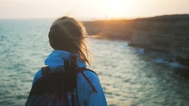Beautiful hiking woman enjoying peaceful sunset on the sea shore. Young girl trekking exploring nature, summer vacation trip. Travel details: wind blowing hair, trekking sticks. Sea waves at sunset.