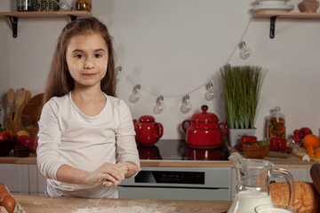 Beautiful little girl dressed in a white blouse is making a dough for baking a bread at a kitchen.