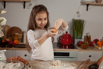 Beautiful little girl dressed in a white blouse is making a dough for baking a bread at a kitchen.