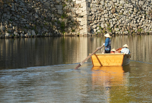 A wooden boat, seen from behind, is controlled by a paddle, and is floating towards a wall made of stones. Warm afternoon light. A lot of negative space for text.