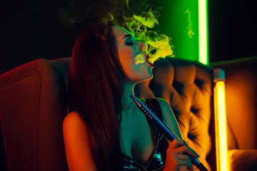 Sexy brunette model is smoking a hookah exhaling a smoke at a luxury night club.