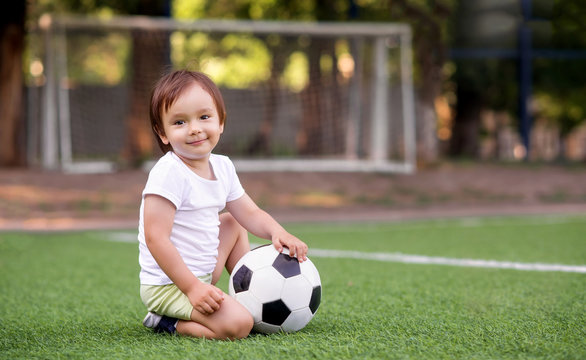 Little toddler boy in sports uniform sitting with soccer ball at football field outdoors in summer day. Goalposts (soccer nets) in background. Kid sports and active childhood concept. Copy space