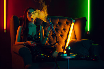 Sexy brunette model is smoking a hookah exhaling a smoke at a luxury night club. - obrazy, fototapety, plakaty