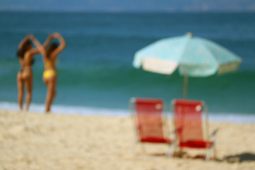 Blurred image of a pair of red beach chairs and light blue parasol on the sandy beach against vivid blue ocean
