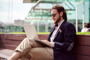 Photo on side of young businessman with laptop sitting on wooden bench in city center on summer day
