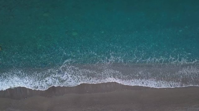 Aerial top view turquoise waves break on empty pebble beach. Pure calm sea from bird's eye view, drone camera locked above blue ocean waves reaching shore and splashing