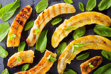 Halves of grilled bananas on a black kitchen board and shale stone. Baked banana dessert with mint on black background. Asian healthy food. Close up view © Lazartivan