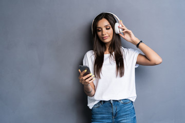 Smiling woman listening music in headphones and using smartphone over gray background.