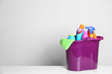 Plastic bucket with different cleaning products on table against light background. Space for text