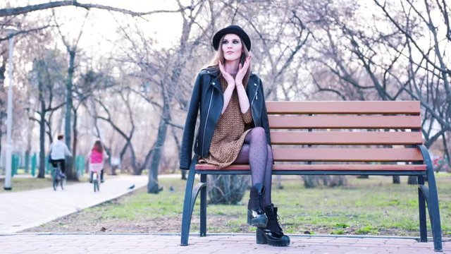 Attractive young woman in fashion dress, black hat, ankle boots and leather jacket sitting alone on the bench in park in warm spring day. Action. Urban modern style