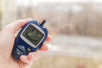 Man holding glucometer with test strip with smile on the monitor on blurred background. Blood glucose meter. Closeup, selective focus