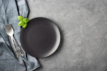 Table setting. Black plate, cutlery and napkin over gray concrete background.
