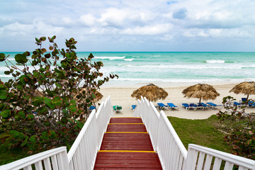Beautiful wooden ladder to the ocean beach. Beach with straw umbrellas and sun-loungers. Cloudy sky and blue water