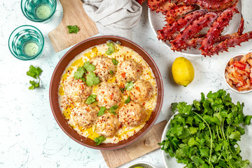 CRABMEATBALLS crab meatballs in white creamy sauce in red pan, whole king crab, cilantro, lemon and white wine on white background