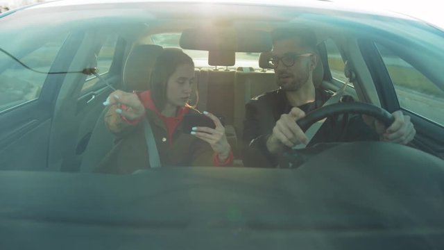 Girlfriend and guy in spectacles sitting in car. Girl telling where to go, pointing finger. Young woman holding smartphone in hands, using navigator app