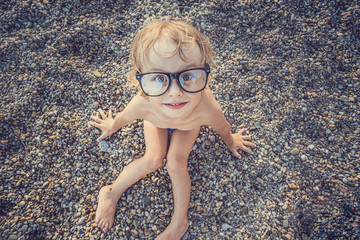 Cute funny little boy in big glasses sunbathing on the pebble beach