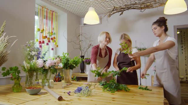 Medium shot of middle-aged blonde female florist at work at flower shop instructing two young trainees on how to stem and strip decorative greenery, working together through bunch of salal tips