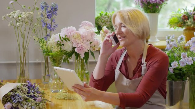 Panning waist-up shot of middle-aged lady florist at work in colorful flower shop, sitting at wooden counter with tablet and talking to customer on mobile phone, with plenty of bouquets everywhere